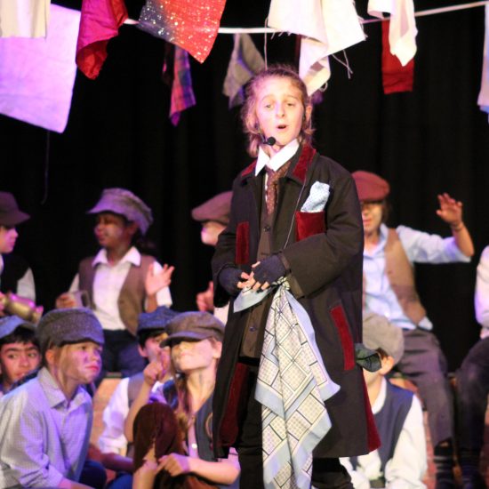 A child singing on the stage with additional actors behind him